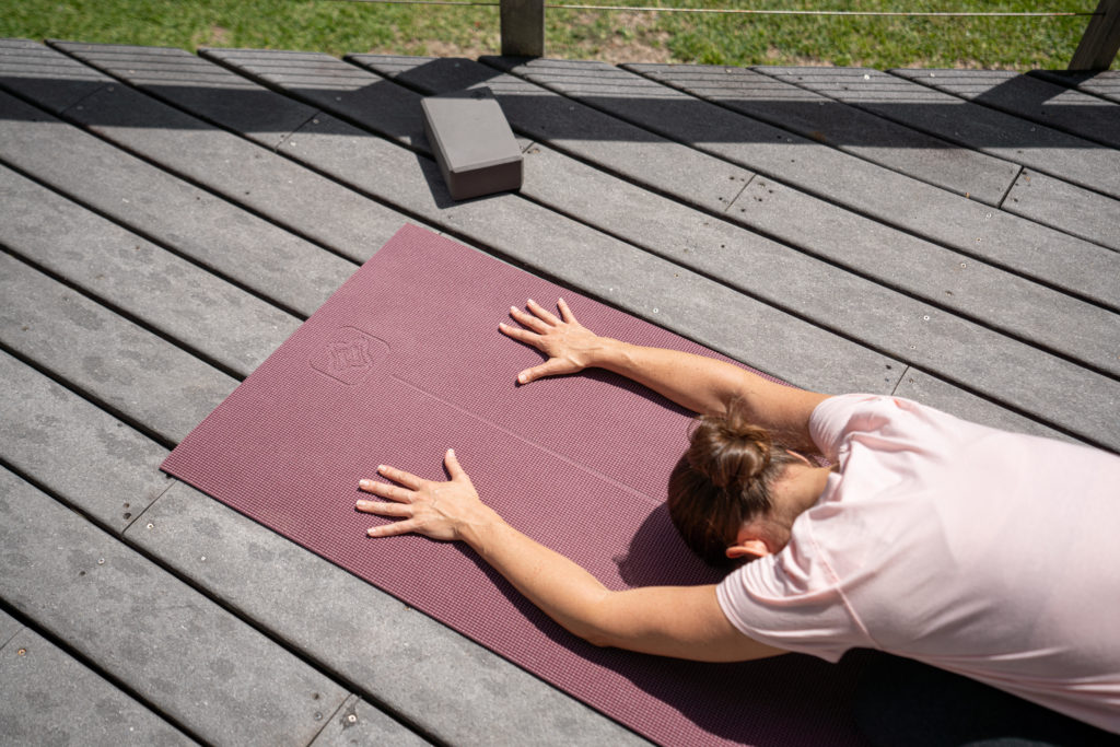 Osoba w Pozycji Dziecka (Balasana) na wysokiej jakości macie do jogi, model DOMYOS-TAPIS-YOGA-DOUX-8MM-PE20, leżącej na drewnianym tarasie. Obok widoczna kostka do jogi.